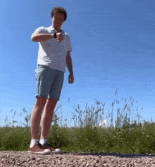 a man in a white shirt and blue shorts is standing on a gravel road looking at his watch .