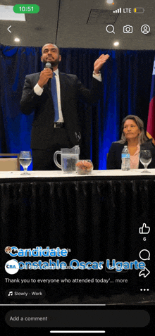 a man in a suit is speaking into a microphone while a woman sits behind him