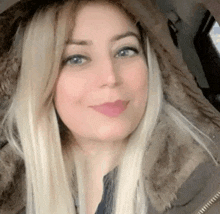 a woman with blonde hair and blue eyes is wearing a fur hooded jacket and smiling for the camera .