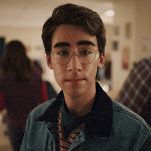 a young man with glasses and a denim jacket looks at the camera