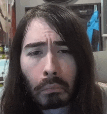 a man with long hair and a beard is looking at the camera with a sad look on his face .