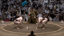 a sumo wrestler is kneeling down in front of a crowd while another wrestler stands in front of a referee