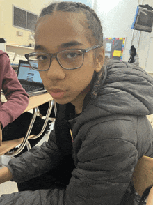 a young girl wearing glasses and a black jacket sits at a desk