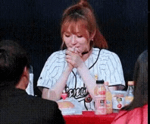 a woman in a baseball jersey is sitting at a table with her hands folded in front of her mouth .