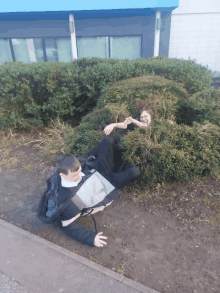 a boy and a girl laying in a bush with a blue building in the background