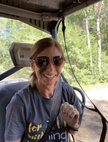 a woman wearing sunglasses and a t-shirt that says " for a wing " smiles while sitting in a vehicle