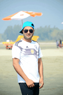 a man wearing a white adidas shirt and a blue hat stands on a beach