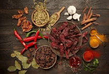 a wooden table topped with a bowl of beef jerky surrounded by spices and herbs .