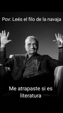 a black and white photo of a man in a suit sitting in a chair with his hands in the air .