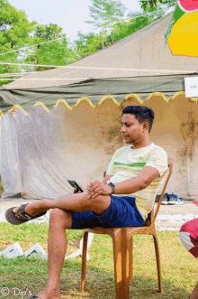 a man sits in a chair with his legs crossed in front of a tent and looks at his phone