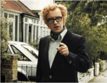 a man with red curly hair and glasses is standing in front of a white car and pointing .