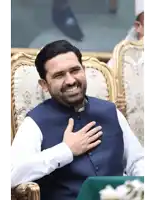 a man wearing a blue vest and white shirt is smiling with his hand on his chest