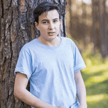 a young man in a light blue t-shirt is leaning against a tree in the woods .