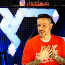 a man in a red shirt with tattoos on his arms is standing in front of a large screen .