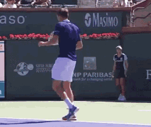 a man in a blue shirt and white shorts is jumping on a tennis court in front of a wall that says masimo