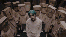 a man with blue hair is sitting in a room surrounded by cardboard boxes with numbers on their heads .