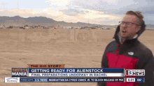 a man in a red and black jacket stands in front of a sign that says the big story getting ready for alienstock