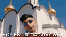 a man wearing sunglasses stands in front of a white building with the words hoy es un dia guay wey above him