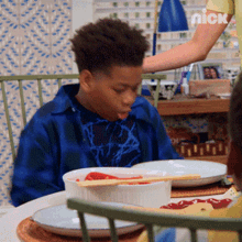 a boy in a blue plaid shirt is sitting at a table with plates and bowls of food and a nick logo in the background