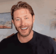 a man with a beard is smiling and wearing a black shirt in a living room .