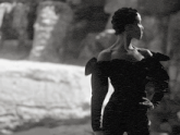a woman in a black off the shoulder dress stands in front of a waterfall