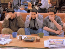 three men sit on a couch covering their faces with their hands while a stack of newspapers sits on a table