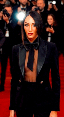 a woman in a tuxedo and bow tie is standing on a red carpet in front of photographers