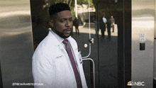 a man in a lab coat and tie stands in front of a glass door with the nbc logo on the bottom