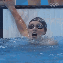 a woman in a swim cap and goggles is swimming in a pool with the olympic rings in the background