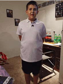 a boy wearing a white shirt and black shorts stands in front of a desk with coca cola and mountain dew