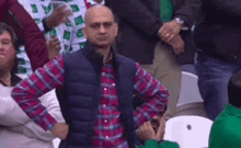 a man in a plaid shirt and vest is sitting in a crowd with his hands on his hips .