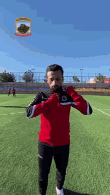 a man in a red and black jacket stands on a soccer field with a logo for a soccer team behind him