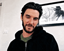 a man with a beard is wearing a black hoodie and smiling in front of a picture on the wall .