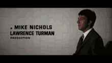 a man in a suit stands in front of a mike nichols lawrence turman production sign