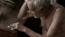 an elderly man without a shirt is sitting at a table with coins in his hands .