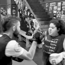 a man and a woman are boxing in a gym . the woman is wearing a shirt that says `` love yourself '' .