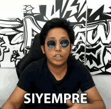 a man wearing sunglasses says siyempre in front of a wall with graffiti