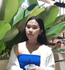 a woman in a blue top is standing in front of a plant holding a cup of coffee .