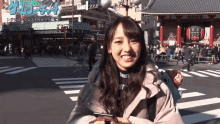 a woman in a pink coat is smiling in front of a crowded street with a sign that says ' dragon quest '