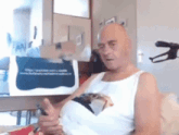 a bald man in a white tank top is sitting on a couch looking at a cell phone