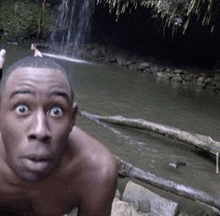 a shirtless man is standing in front of a waterfall and looking at the camera .