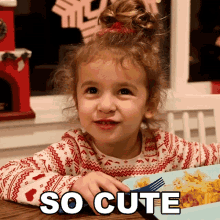 a little girl is sitting at a table with a plate of food and the words so cute above her head