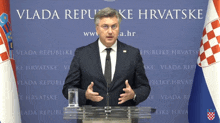 a man in a suit and tie stands at a podium in front of a wall that says vlada republike