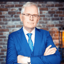 a man wearing a blue suit and tie is standing with his arms crossed in front of a brick wall .