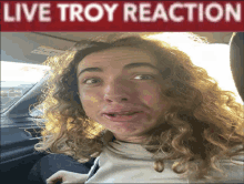 a man with curly hair is sitting in a car under a live troy reaction banner