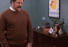 a man in a brown sweater is standing in front of a desk with flags on it