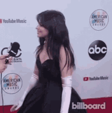 a woman in a black dress and white gloves is standing on a red carpet at a billboard event .