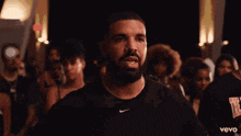 a man with a beard and a nike shirt is standing in front of a crowd of people .