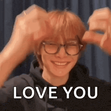 a man wearing glasses and a hoodie is smiling and making a heart with his hands .