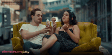 a man and woman are sitting on a yellow couch eating ice cream